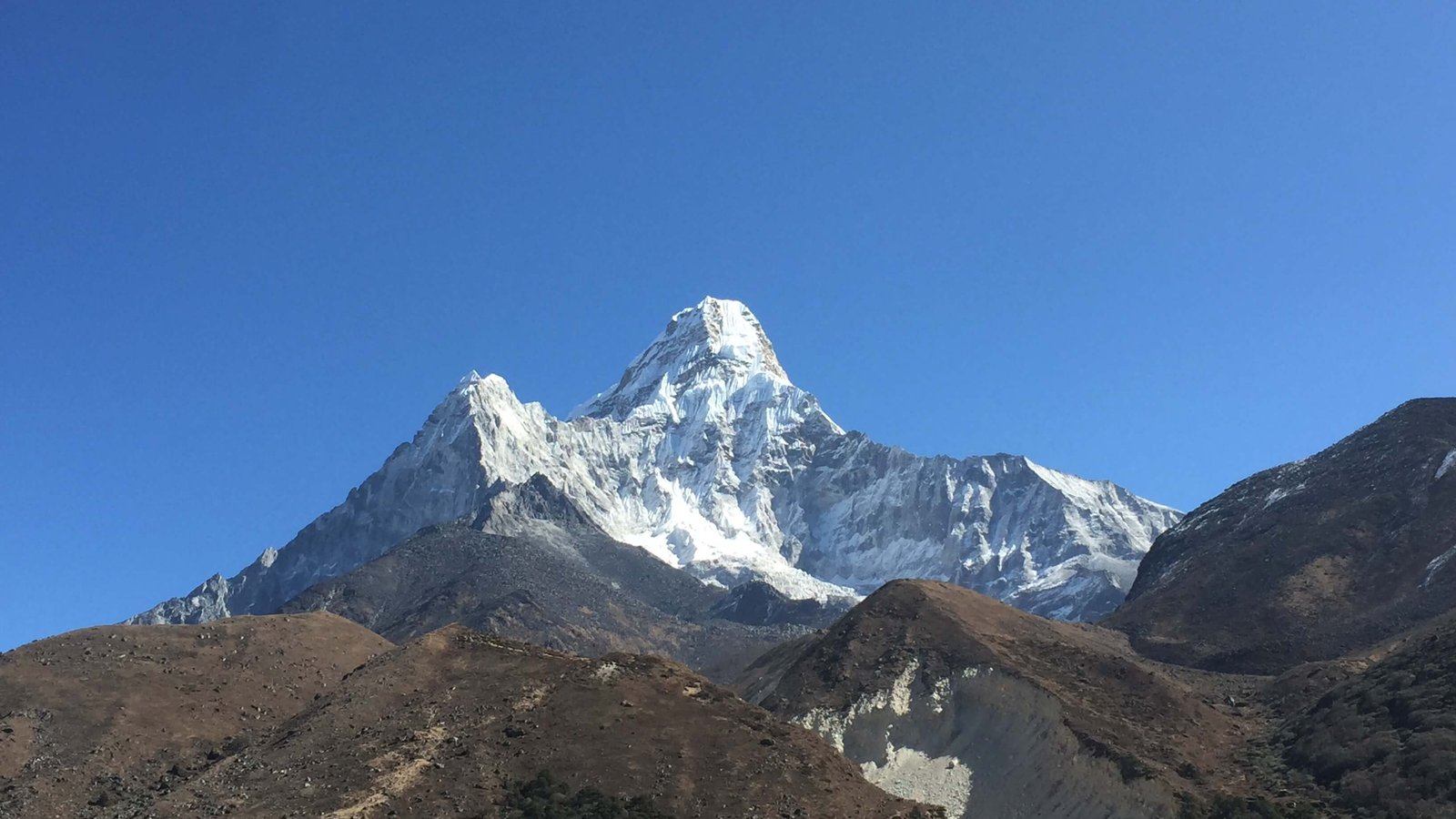 Mount Ama Dablam under clear blue sky, viewed from Everest trekking trail in Nepal with brown ridges and rocky Himalayan terrain in the foreground