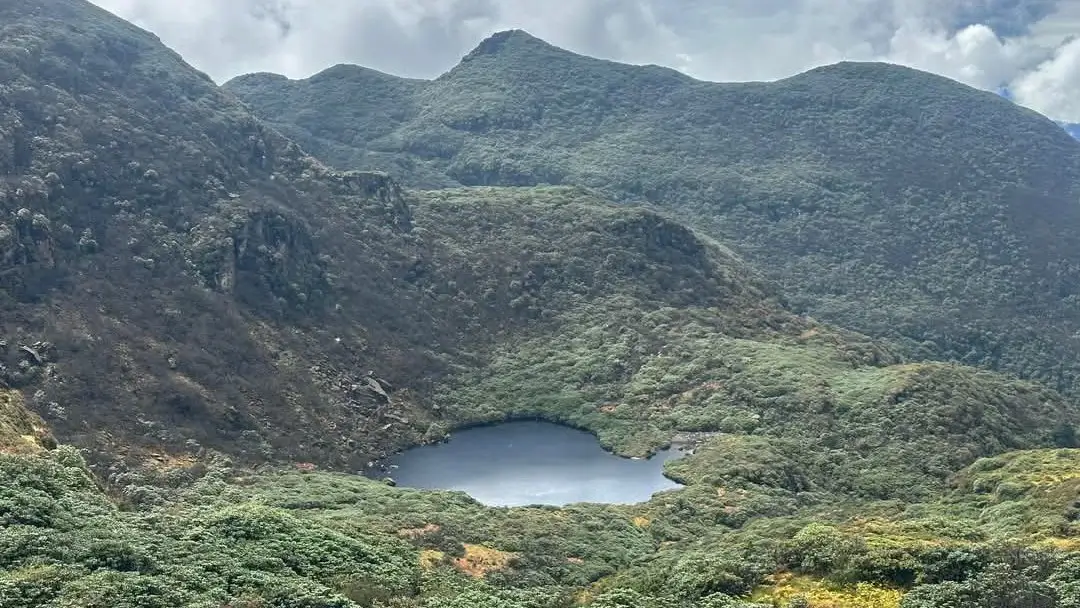 High-altitude mountain lake nestled among green hills and rugged peaks, under a cloudy sky in a remote wilderness area.