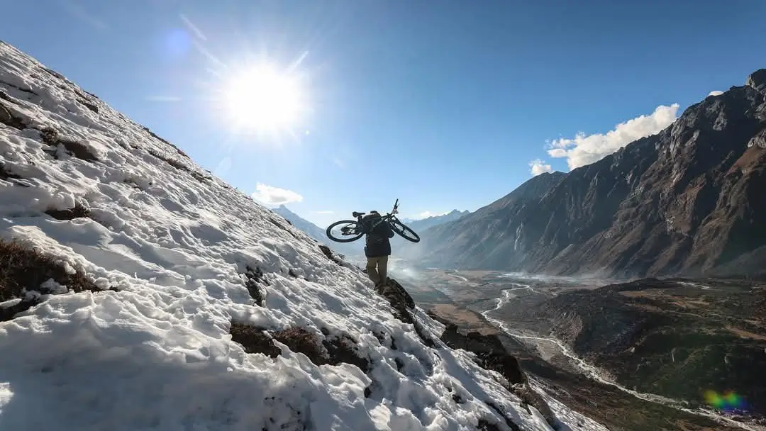 Trekker climbing a steep snow-covered mountain slope with mountain bike under a bright sun, surrounded by Himalayan peaks and a clear blue sky.