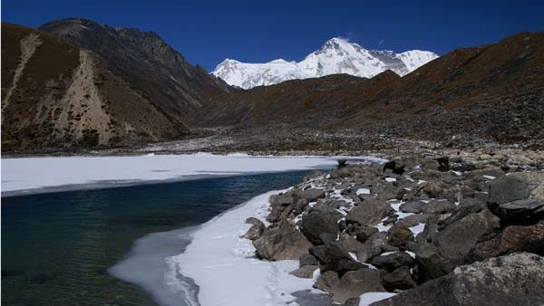 Frozen Gokyo Lake with snow-covered rocky shore and Himalayan peaks in the background on a clear blue sky day in the Everest Region of Nepal